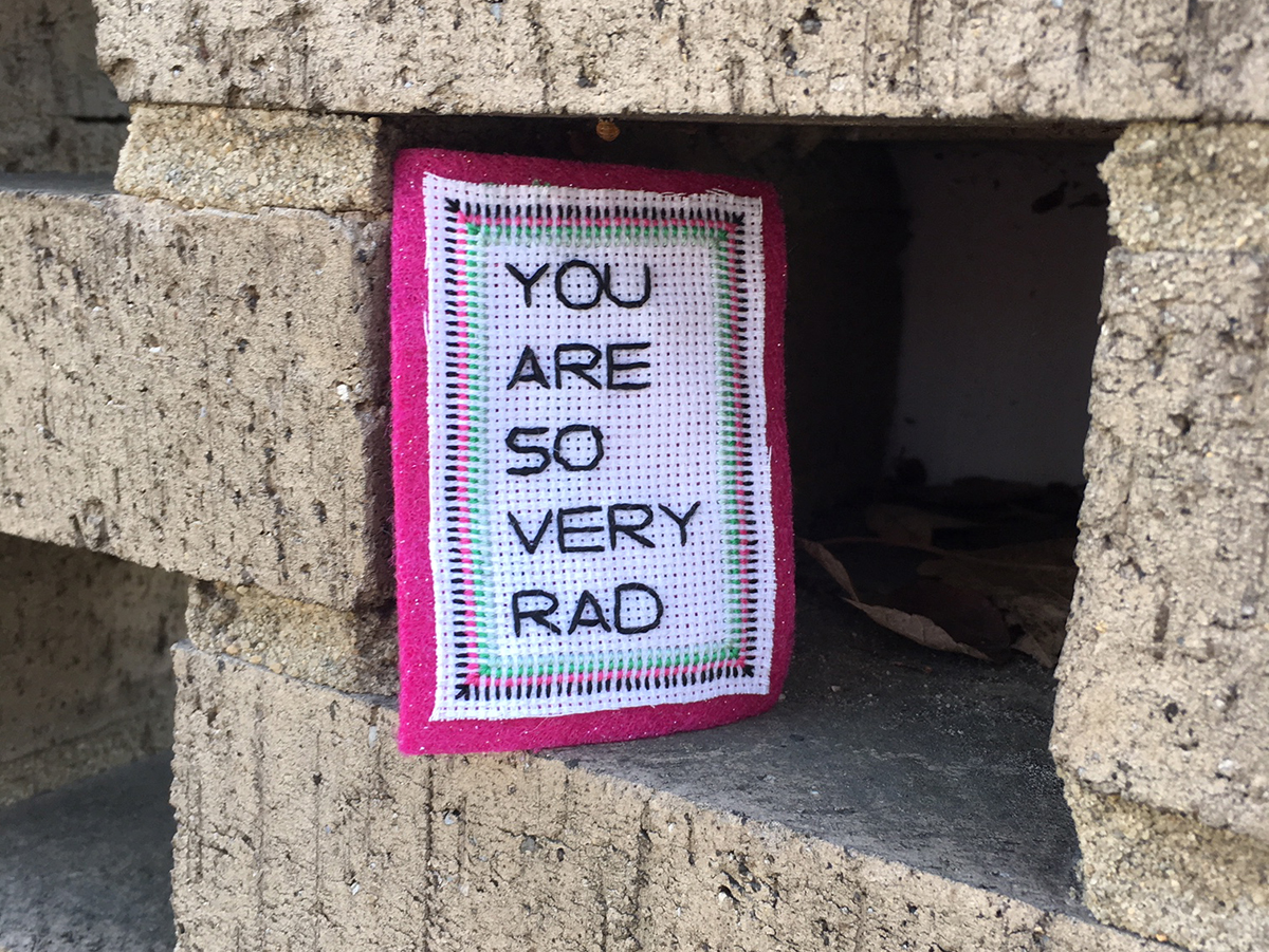 Betsey Greer’s colorful “You are so very rad” stitch placed in between the bricks of a wall.